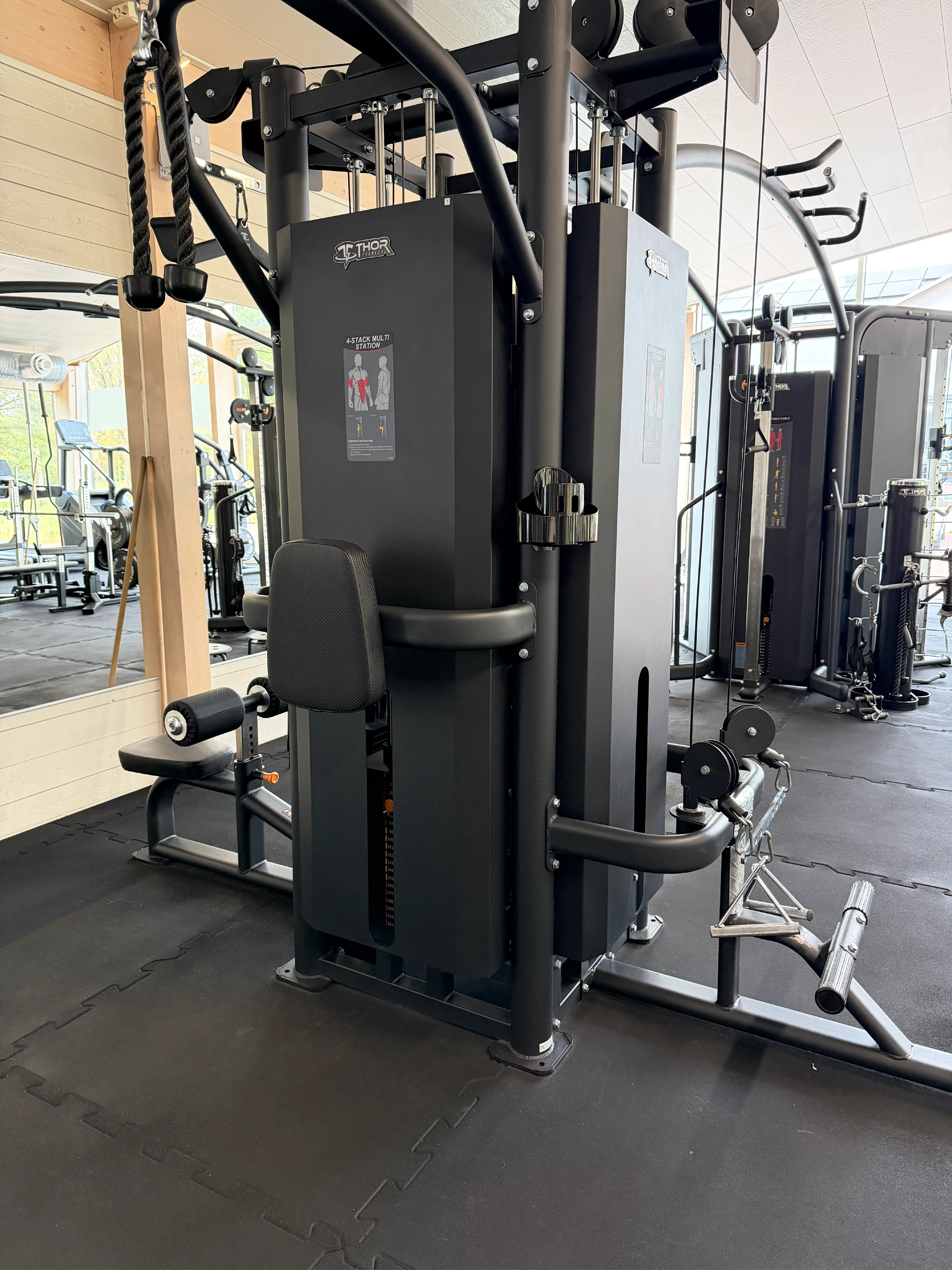 Hälsofabrikens cable cross-maskin i svart, utrustad med flera handtag och viktstationer för mångsidig styrketräning. Maskinen står på svart gummigolv i ett ljust gym med stora fönster.