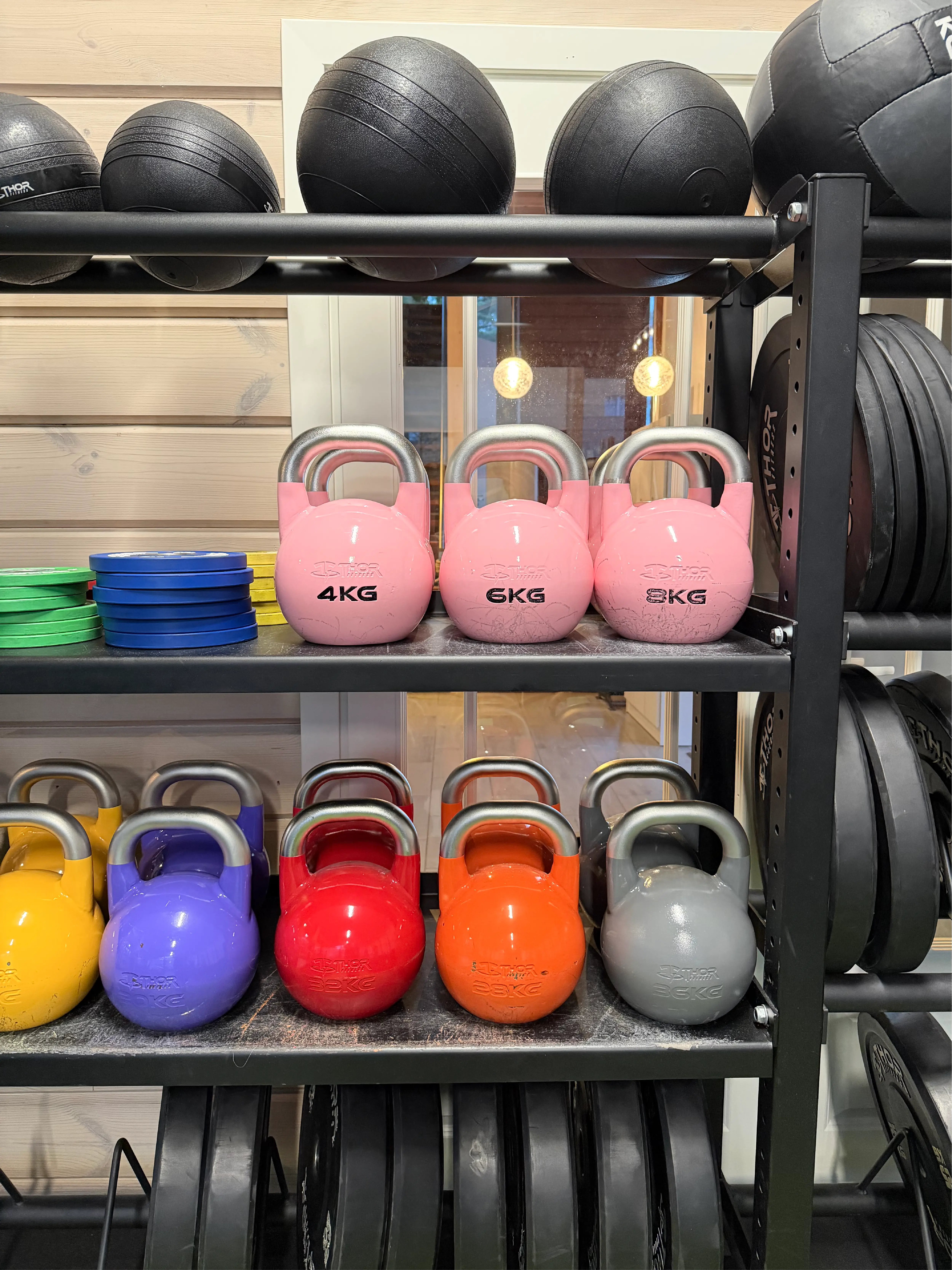 Kettlebells i olika färger och vikter uppställda på hylla tillsammans med viktskivor och bollar