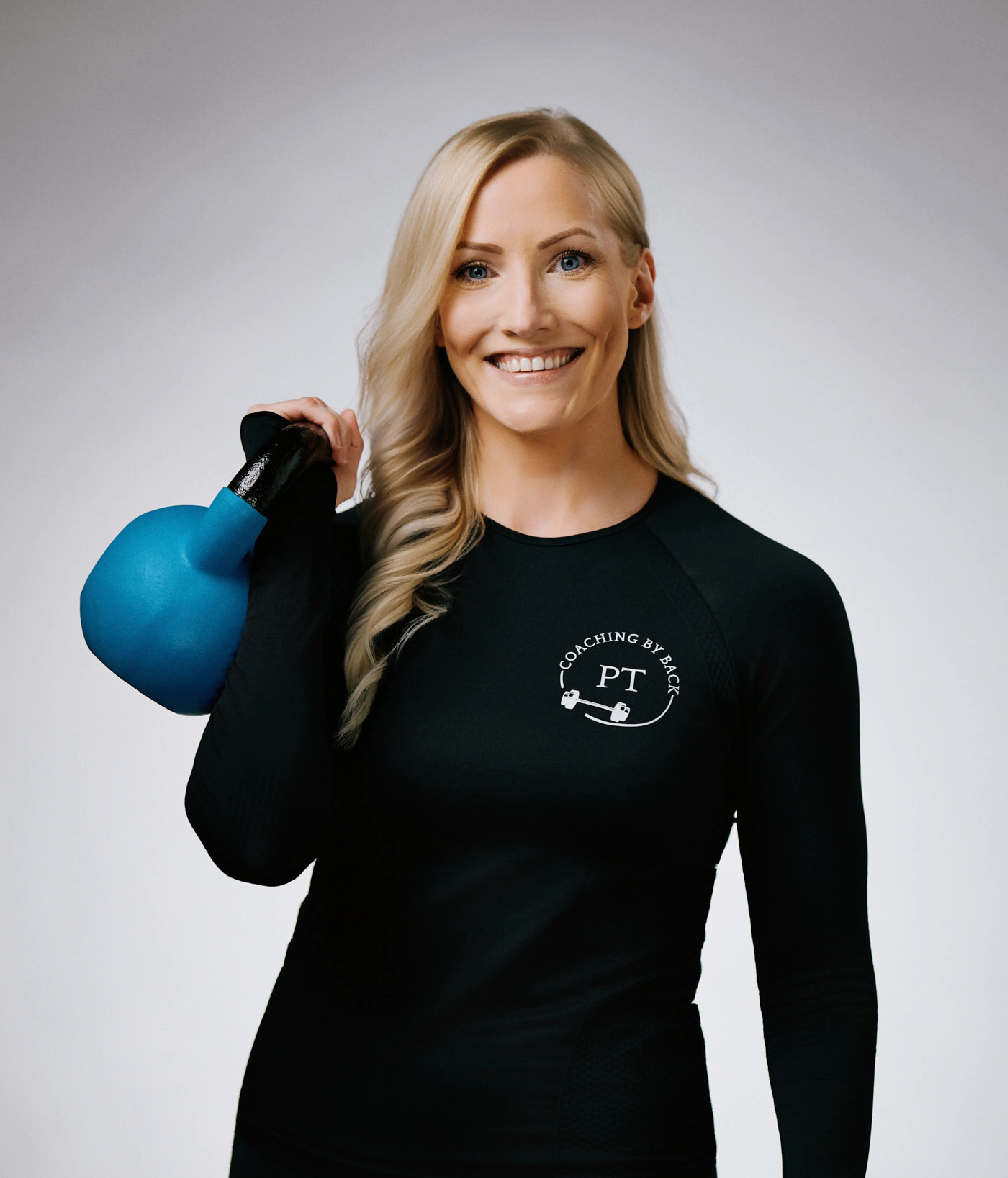 Porträtt av Camilla Back, personlig tränare på Hälsofabriken. Hon ler mot kameran och håller en blå kettlebell vilande på axeln. Klädd i svart träningskläder med logotypen 'Coaching by Back PT'. Bakgrunden är ljus och neutral.