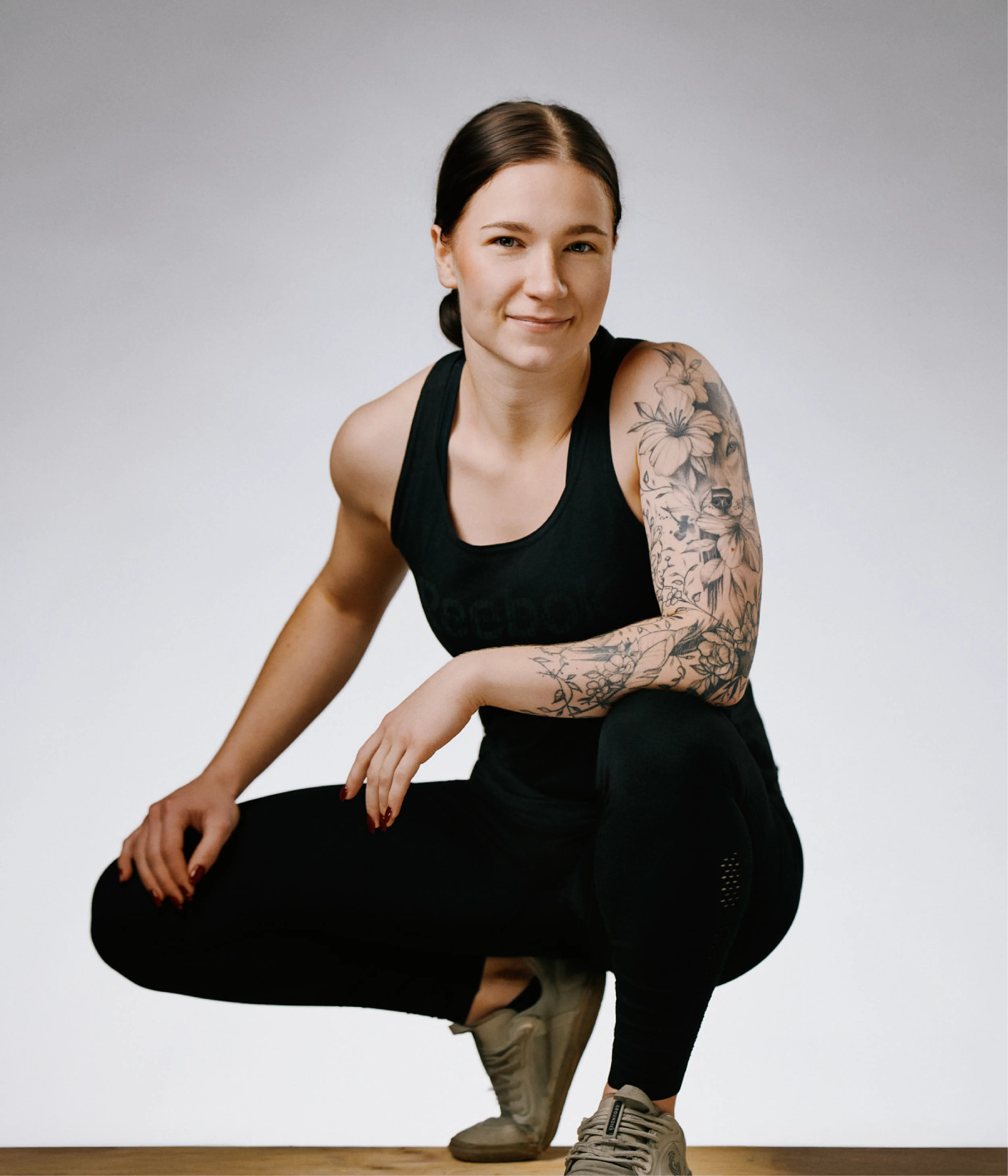 Porträtt av Peppi Nyström, personlig tränare på Hälsofabriken. Hon sitter på huk och ler mot kameran, klädd i svarta träningskläder. Hennes ena arm är täckt av en detaljerad blomtatuering. Bakgrunden är ljus och neutral.